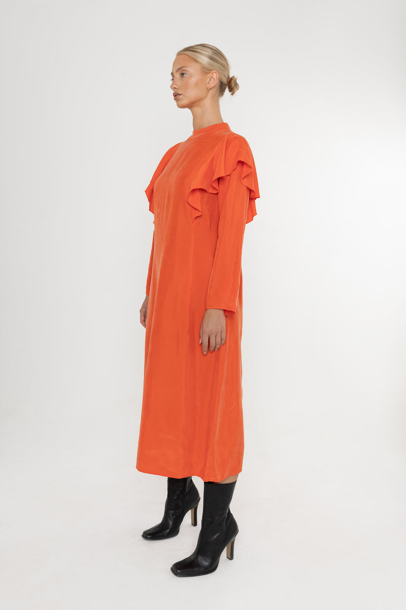 Woman wearing an orange vermillion red dress with ruffled long sleeves and black boots on a white background