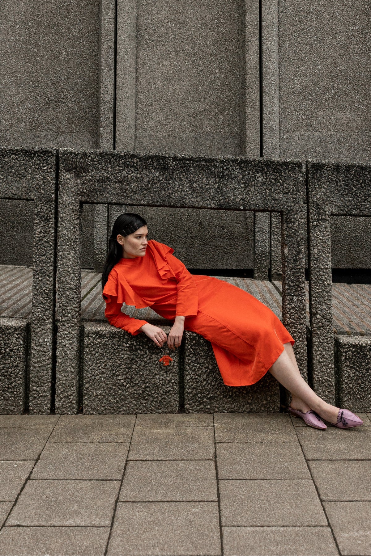 A woman wearing a bright orange-red dress sits on a concrete bench against a textured gray concrete wall.