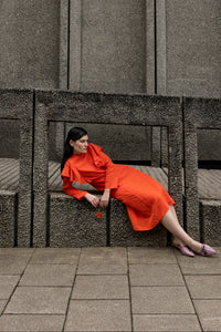 A woman wearing a bright orange-red dress sits on a concrete bench against a textured gray concrete wall.
