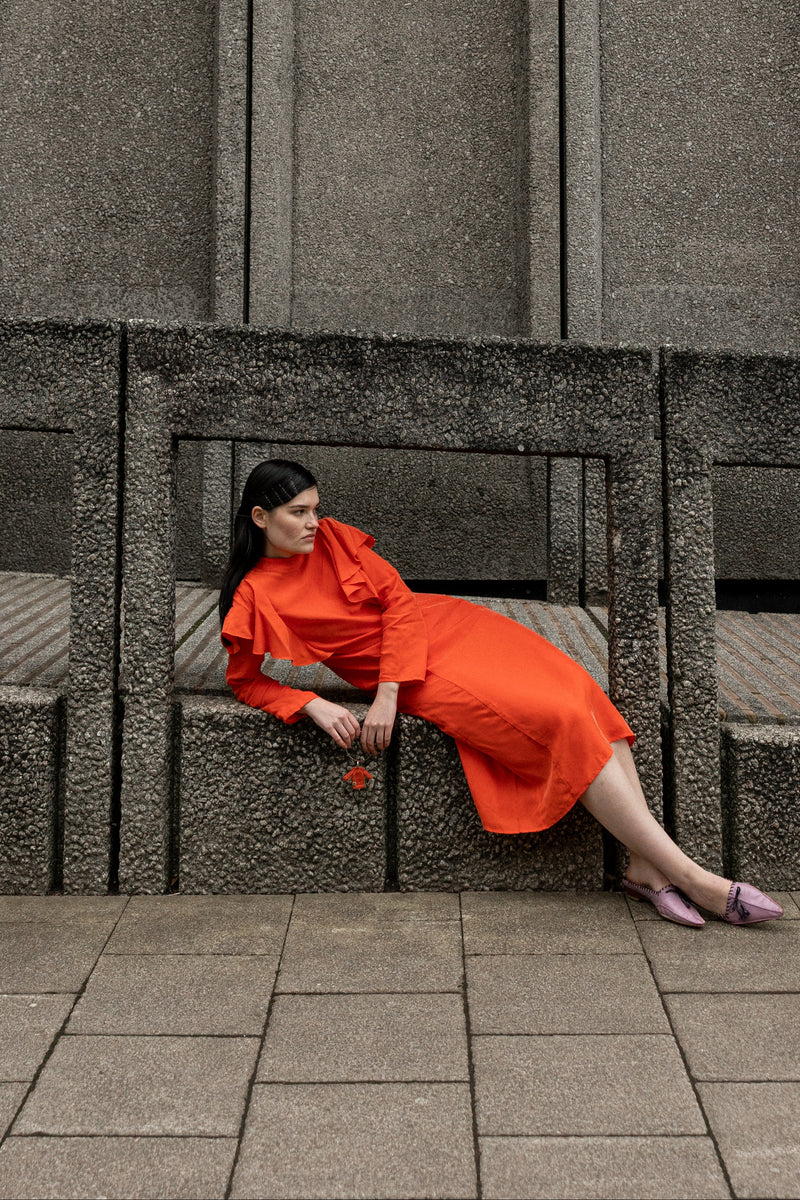 A woman wearing a bright orange-red dress sits on a concrete bench against a textured gray concrete wall.
