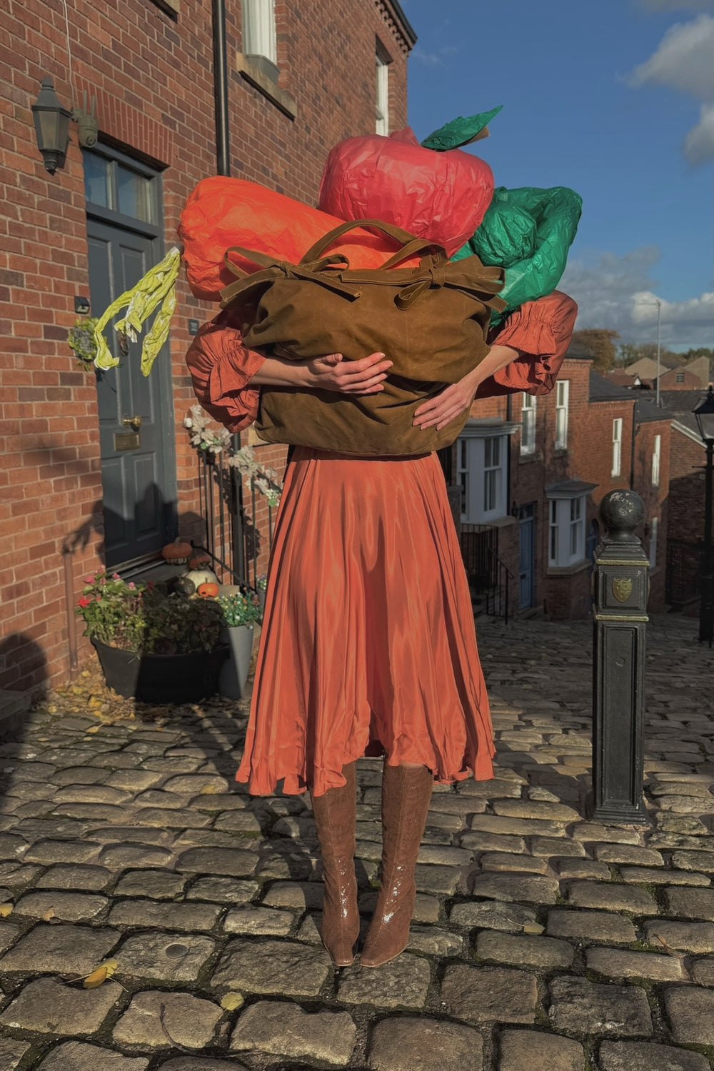 Person in an orange florence Deidei dress carrying oversized produce on a cobblestone street.