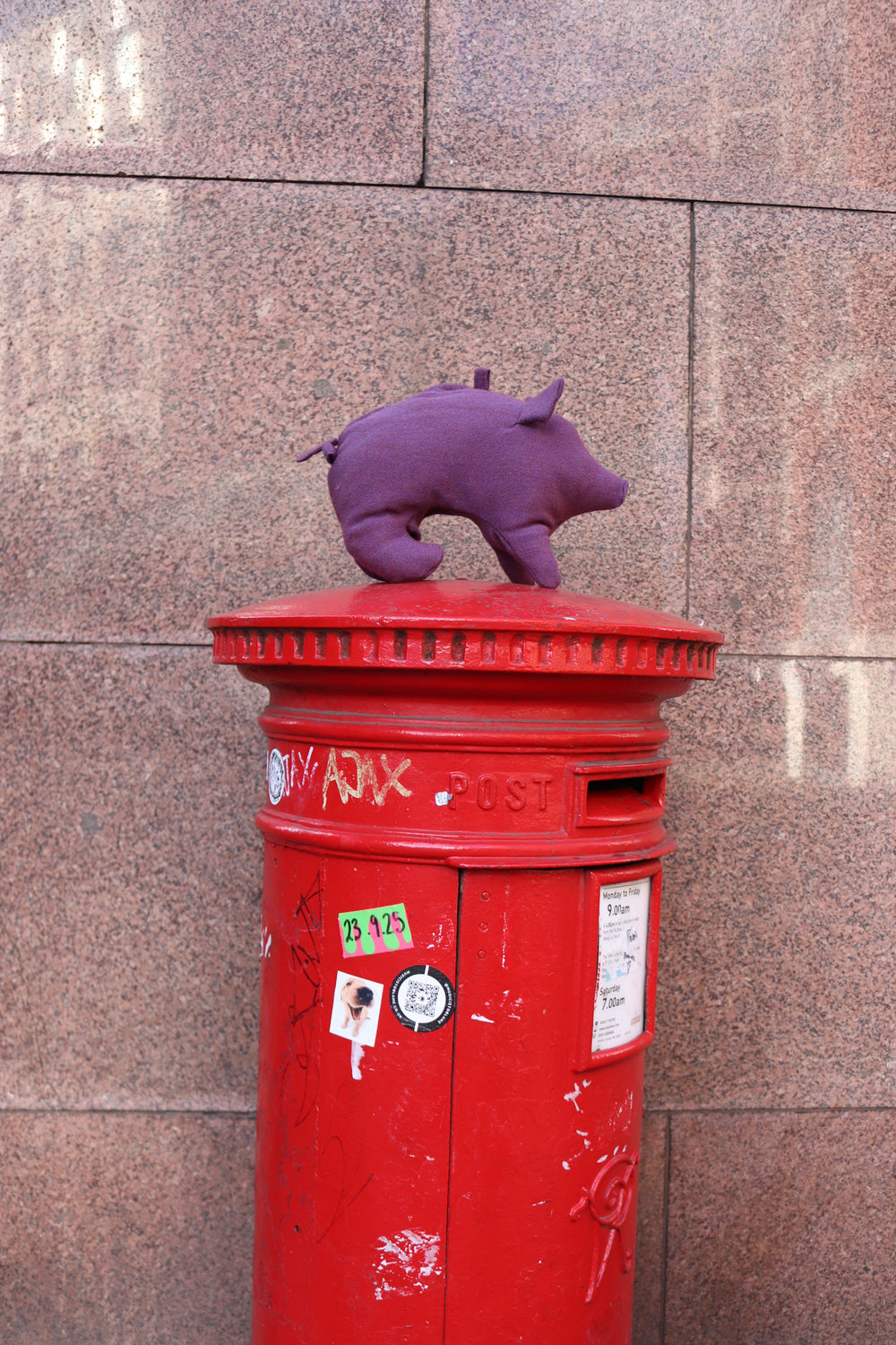 Red postbox with a purple pig plush toy or bag against a tiled wall.